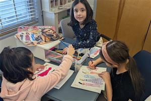 3 school-aged girls coloring fashion designs at a table.